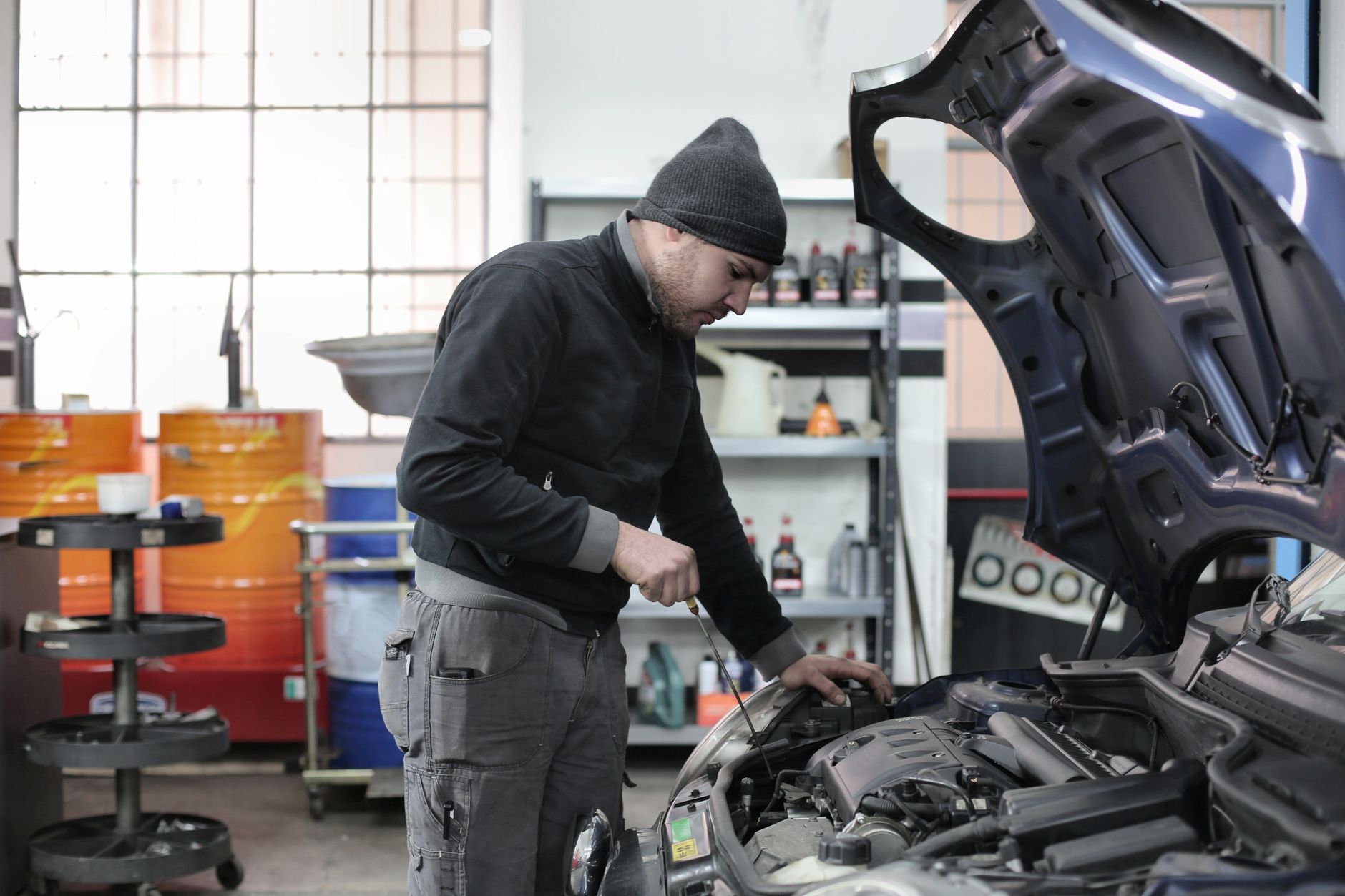 Mechanic in dark hoodie and beanie works under open car hood, using tools in a workshop.